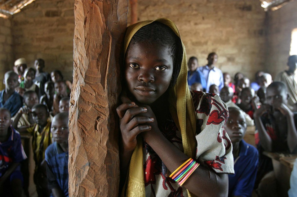 Primo piano di ragazza congolese appoggiata ad un palo di legno, in una stanza piena di persone